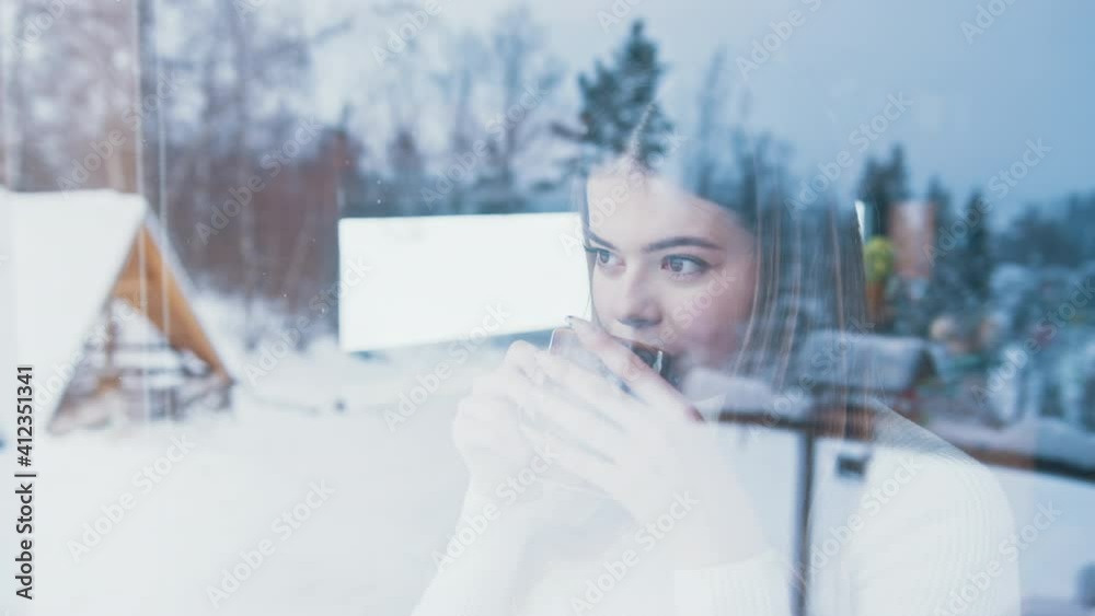 Young caucasian woman drinking hot beverage and looking through the window at snow covered houses. High quality 4k footage