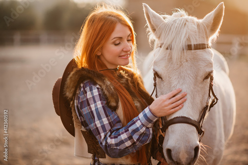 A young cowgirl woman stroking a horse in the sunset at her ranch. Flare effect. © MZaitsev