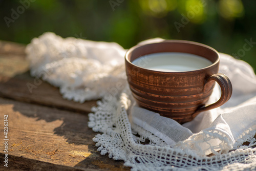 Pottery cup with milk (kefir, yogurt, sour cream, kumis), lace tablecloth, wooden table. Outdoor picnic, breakfast, brunch, refreshments. Soft focus photo