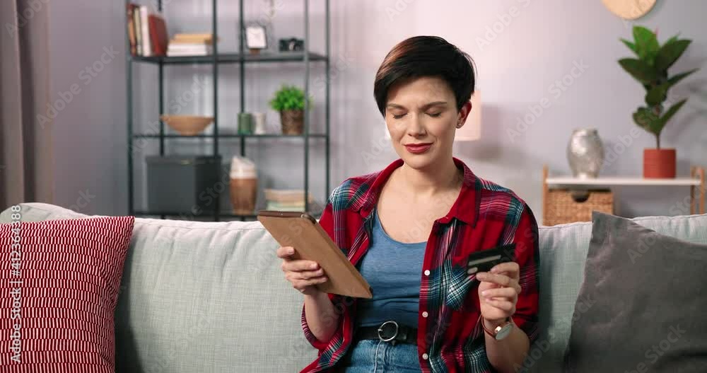 Great shopping. A beautiful happy caucasian brunette girl or young woman sitting at the sofa and using tablet while shopping online using her credit card. Woman typing credit card number at the tablet Great shopping. A beautiful happy caucasian brunette girl or young woman sitting at the sofa and using tablet while shopping online using her credit card. Woman typing credit card number at the tablet