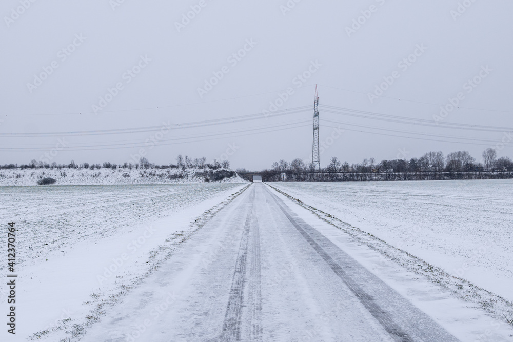 Outdoor scenery diminishing perspective view on street along land and field covered with thick layer snow and background of cloudy overcast sky in countryside in Germany during winter season.