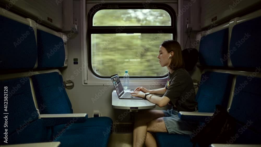Beautiful young woman in casual clothes working on a laptop at a table in a train and communicates on wireless headphones. Female freelancer working on laptop while traveling by train.