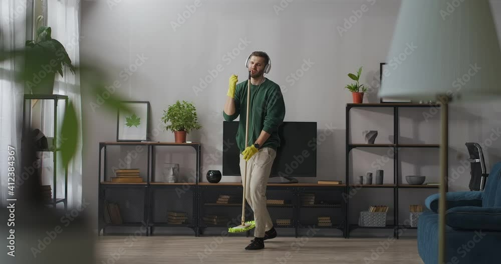 adult handsome man is singing into mop during cleaning in apartment, listening to music in headphones, fun and amusement during home clean-up adult handsome man is singing into mop during cleaning in apartment, listening to music in headphones, fun and amusement during home clean-up