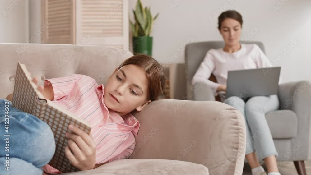 Lockdown of teenage Caucasian girl lying on couch at home and reading interesting book while her mom sitting on armchair with laptop on her knees on background Lockdown of teenage Caucasian girl lying on couch at home and reading interesting book while her mom sitting on armchair with laptop on her knees on background