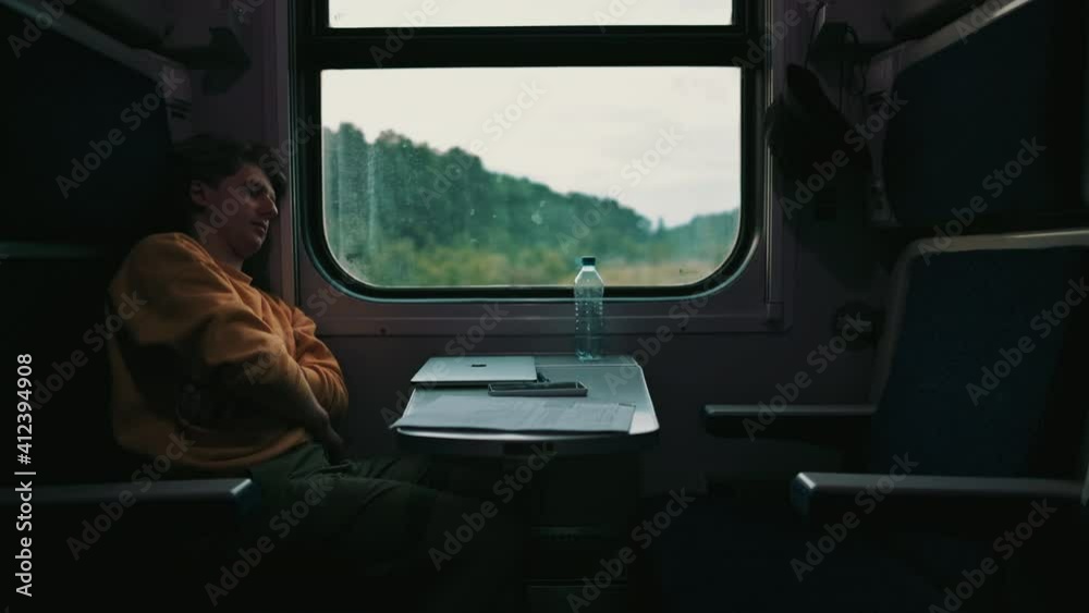 Sleepy young man rides a train by the window in the evening, closes his laptop and falls asleep. Freelancer is tired of working on the road.