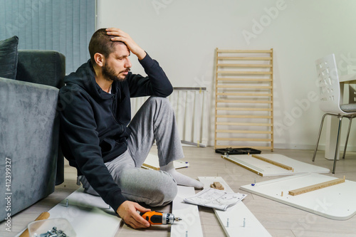 One Man with beard alone Putting Together Self Assembly Furniture at Home holding electric screwdriver looking the instructions frustrated desperate hold head - DIY concept real people copy space photo