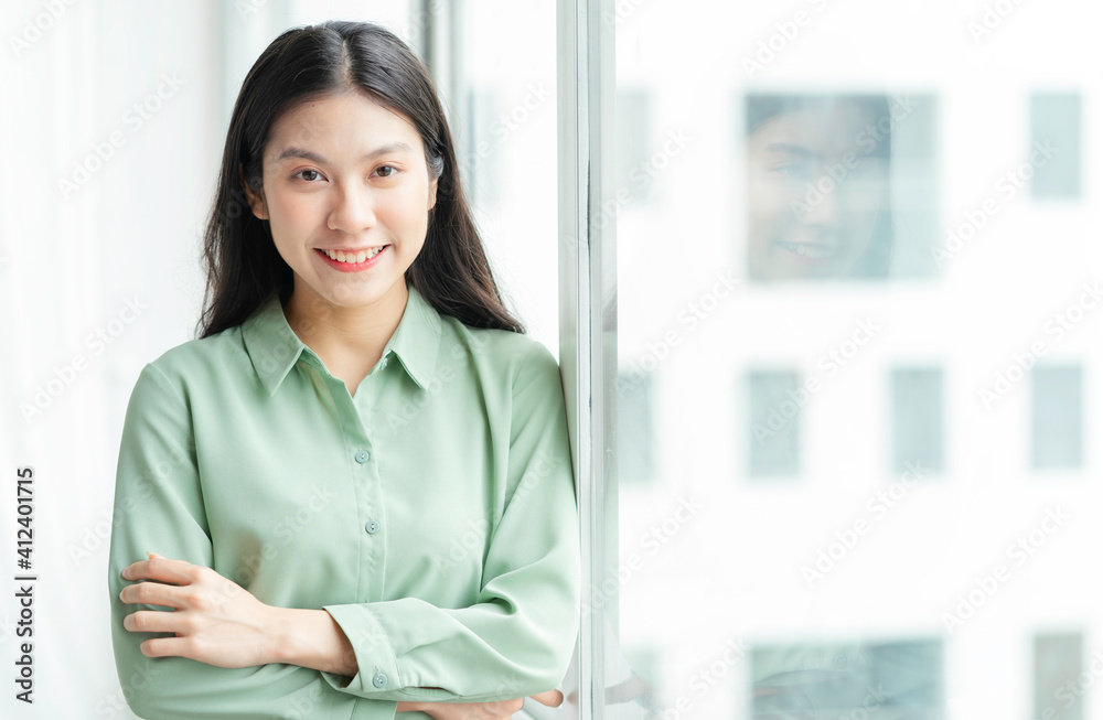 Portrait of young Asian businesswoman standing by the glass door with cheerful expression Portrait of young Asian businesswoman standing by the glass door with cheerful expression