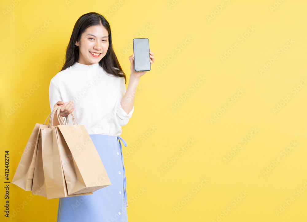 Young girl holding shopping bag with happy expression is using phone on yellow background Young girl holding shopping bag with happy expression is using phone on yellow background