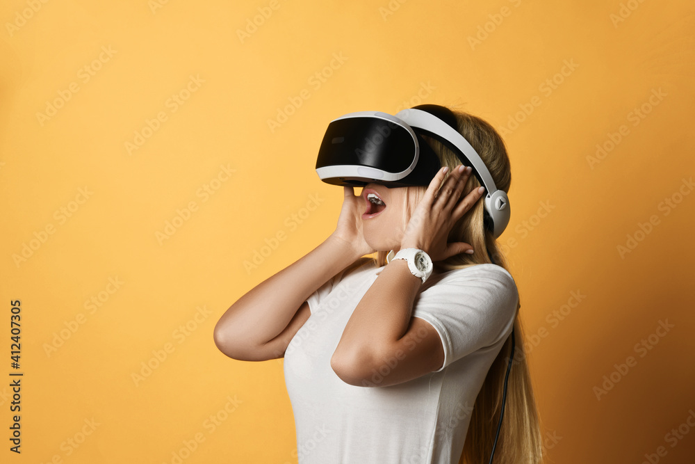 Portrait of young blonde woman in white t-shirt and VR glasses amazed by modern augmented virtual reality technology holding hands at her head. Modern cyber technologies concept Portrait of young blonde woman in white t-shirt and VR glasses amazed by modern augmented virtual reality technology holding hands at her head. Modern cyber technologies concept