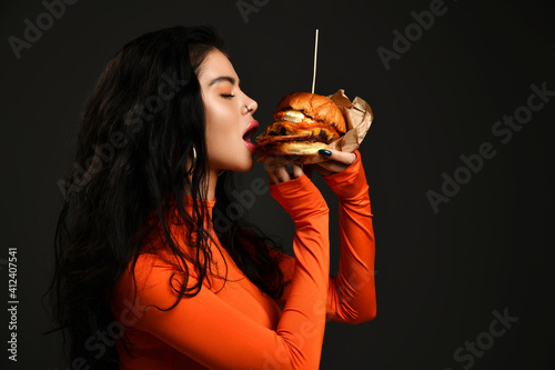 Valokuva Portrait of hot sexy brunette woman with pouty lips in tight red clothes eating biting fresh big tasty craft burger with her eyes closed over black background