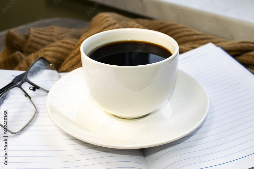 On the saucer is a white cup of coffee. A notebook and glasses for vision on a warm cozy sweater. On the saucer is a white cup of coffee. A notebook and glasses for vision on a warm cozy sweater.