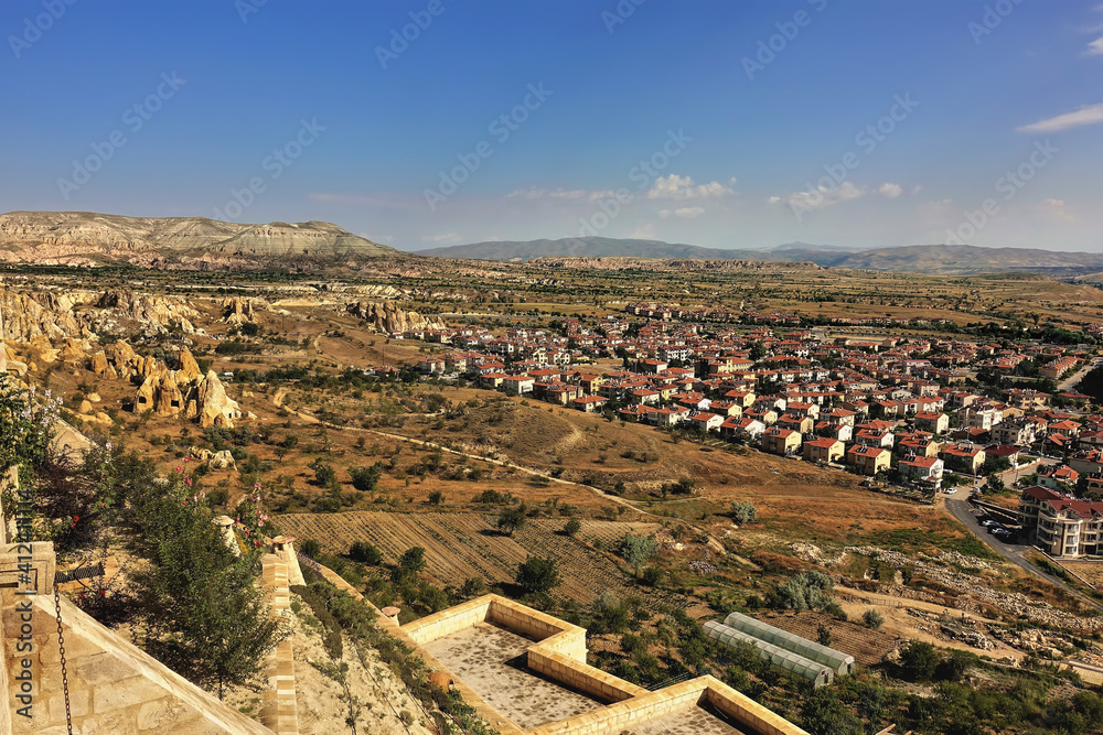 Panorama of the Cappadocia Valley at golden hour. A village is visible on the plain: several houses with red roofs. Around the peaked rocks with caves, a mountain with a flat top. Blue sky. Turkey