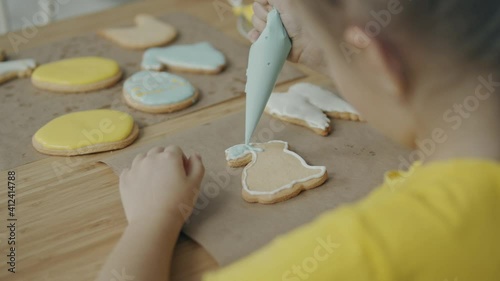 A blonde girl in a yellow T-shirt sits at a table at home and decorates cookies with icing. Family concept. Easter and Easter cookies concept.