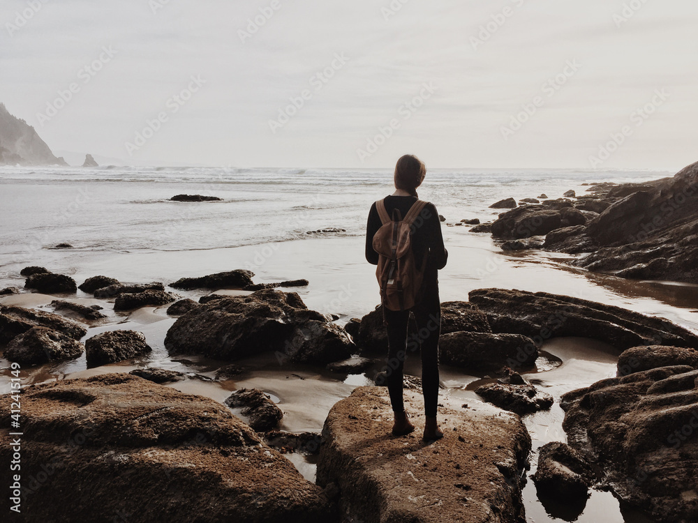 Girl wearing a backpack standing on rocks near the ocean shore Girl wearing a backpack standing on rocks near the ocean shore