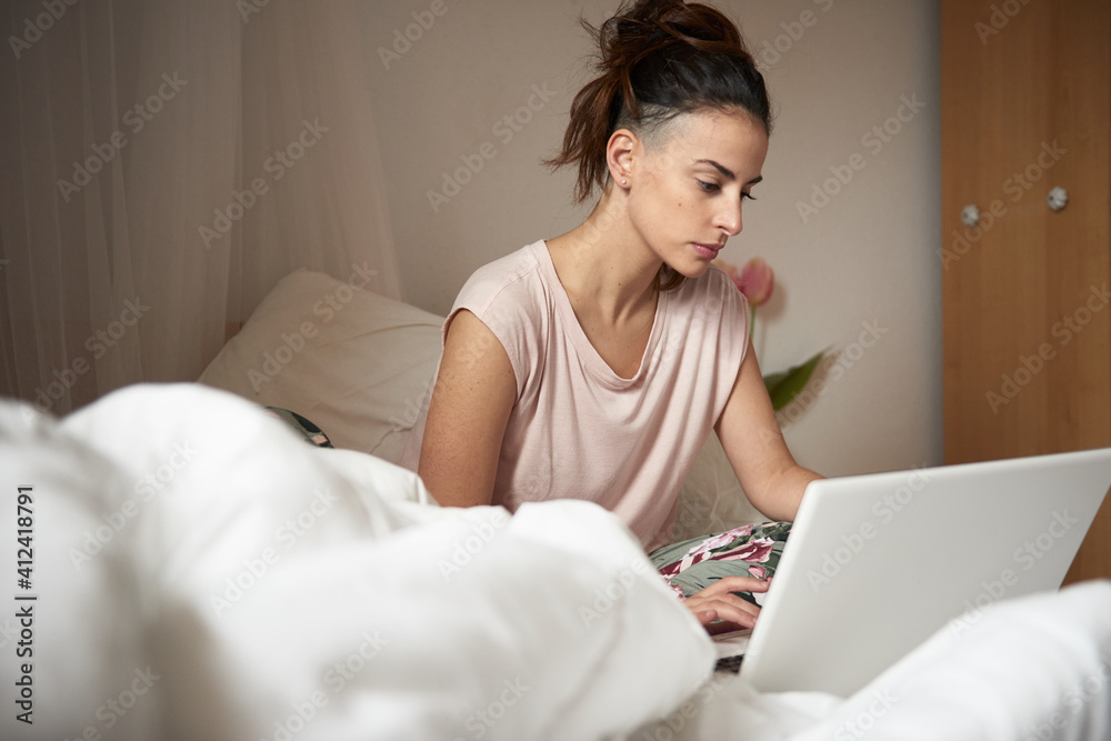 Brunette wearing home clothes sitting on bed with laptop Brunette wearing home clothes sitting on bed with laptop