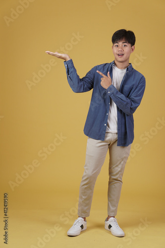 portrait of happy smiling young asian man pointing finger to empty open hand isolated on yellow background