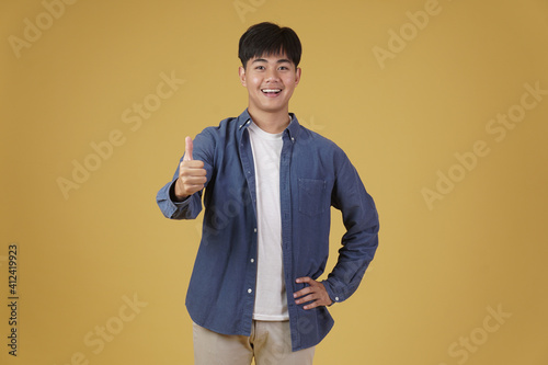 portrait of smiling positive young asian man dressed casually with thumb up gesture approving expression isolated on yellow background