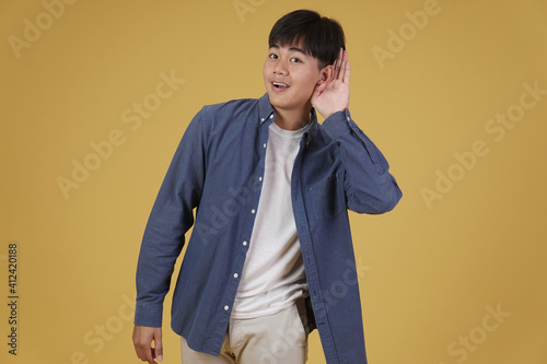 portrait of cheerful happy rejoicing young asian man dressed casually putting hand on ear listening to something isolated on yellow background
