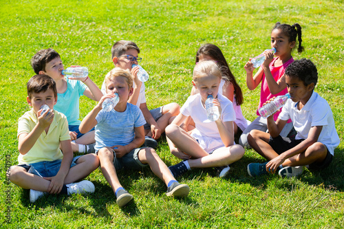 Group of happy kids drinking water and sitting on grass in park outdoors