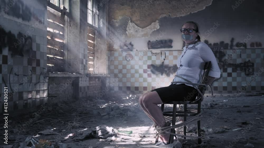 Frightened woman in captivity inspects room while sitting tied to chair.