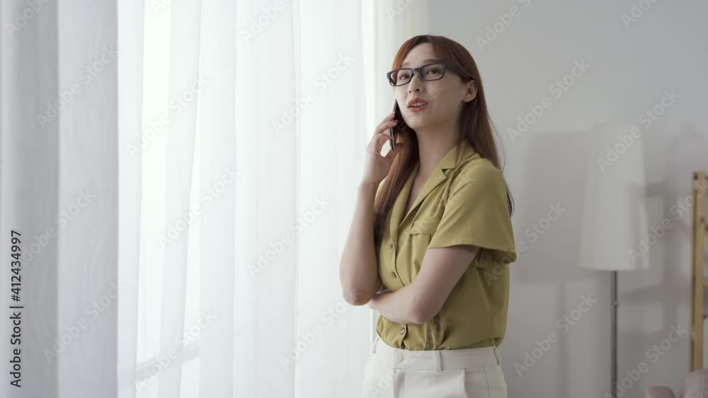 asian female entrepreneur standing by window is flicking hair while having business talk on phone. asian female entrepreneur standing by window is flicking hair while having business talk on phone.