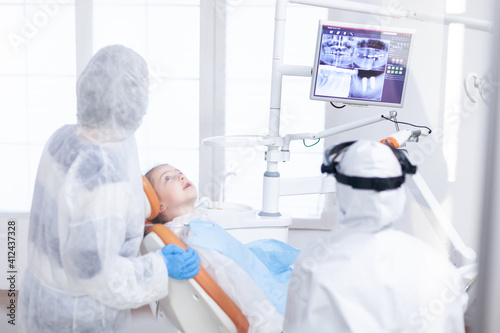 Little girl in ppe looking at her mother in the course of dental examination. Stomatolog in protectie suit for coroanvirus as safety precaution looking at digital child teeth x-ray during consultation