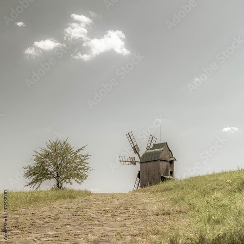Old wodden windmill on a green meadow, field. Lonely green tree in the wind. Blue sky with white clouds. Old photo.