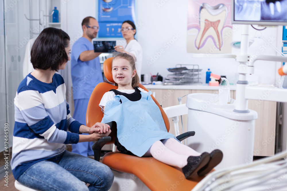 Little girl missing milk tooth wearing dental bib discussing with parent at dentist consultation. Child with her mother during teeth check up with stomatolog sitting on chair. Little girl missing milk tooth wearing dental bib discussing with parent at dentist consultation. Child with her mother during teeth check up with stomatolog sitting on chair.