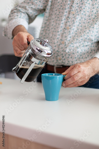 Retired man holding blue cup and french press on kitchen table pouring hot aromatic drink from a coffee machine during breakfast. Elderly person in the morning drinking fresh brown espresso from mug