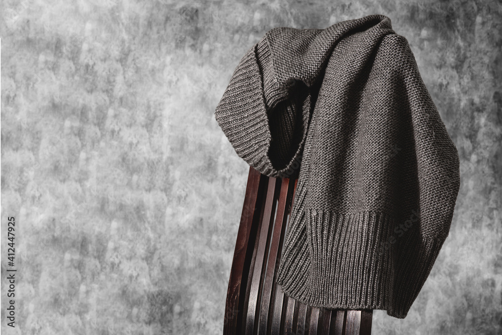 Knitted gray sweater on the back of wooden chair against gray concrete color wall. Minimal style. Copy space.
