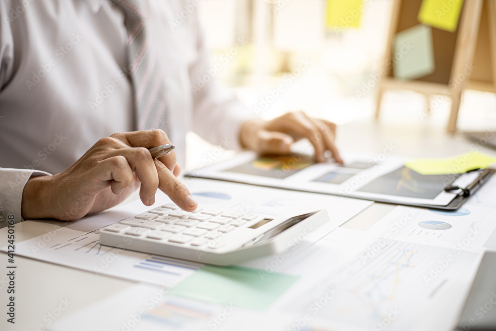 A businessman is using a white calculator to calculate financial numbers, he is sitting in his private office, the businessman examines the financial data from the corporate finance chart. A businessman is using a white calculator to calculate financial numbers, he is sitting in his private office, the businessman examines the financial data from the corporate finance chart.