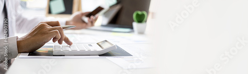 Businessman is using a calculator to calculate company financial figures from earnings papers, a businessman sitting in his office where the company financial chart is placed. Banner with copy space.