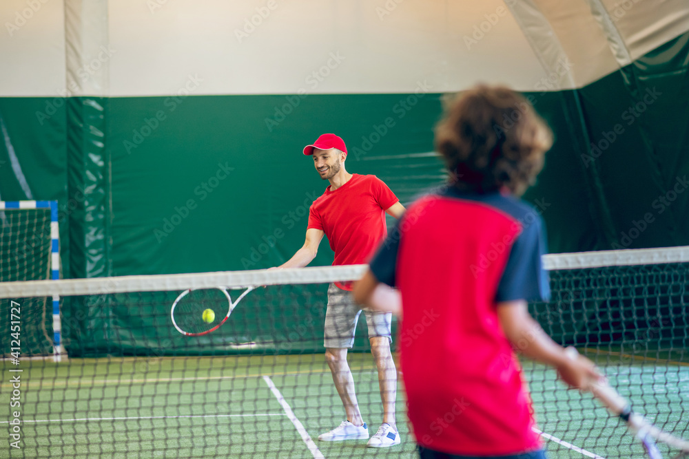 Coach in red clothes and cap playing tennis with a boy