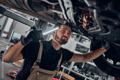 Portrait shot of a handsome mechanic working on a vehicle in a car service. Professional repairman is wearing gloves and using a lamp underneath the car.