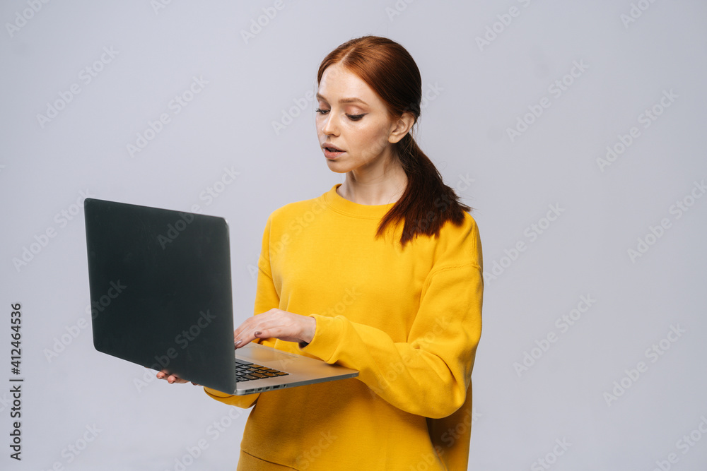 Serious young woman student holding laptop computer and typing on isolated gray background. Pretty redhead lady model emotionally showing facial expressions in studio, copy space. Serious young woman student holding laptop computer and typing on isolated gray background. Pretty redhead lady model emotionally showing facial expressions in studio, copy space.