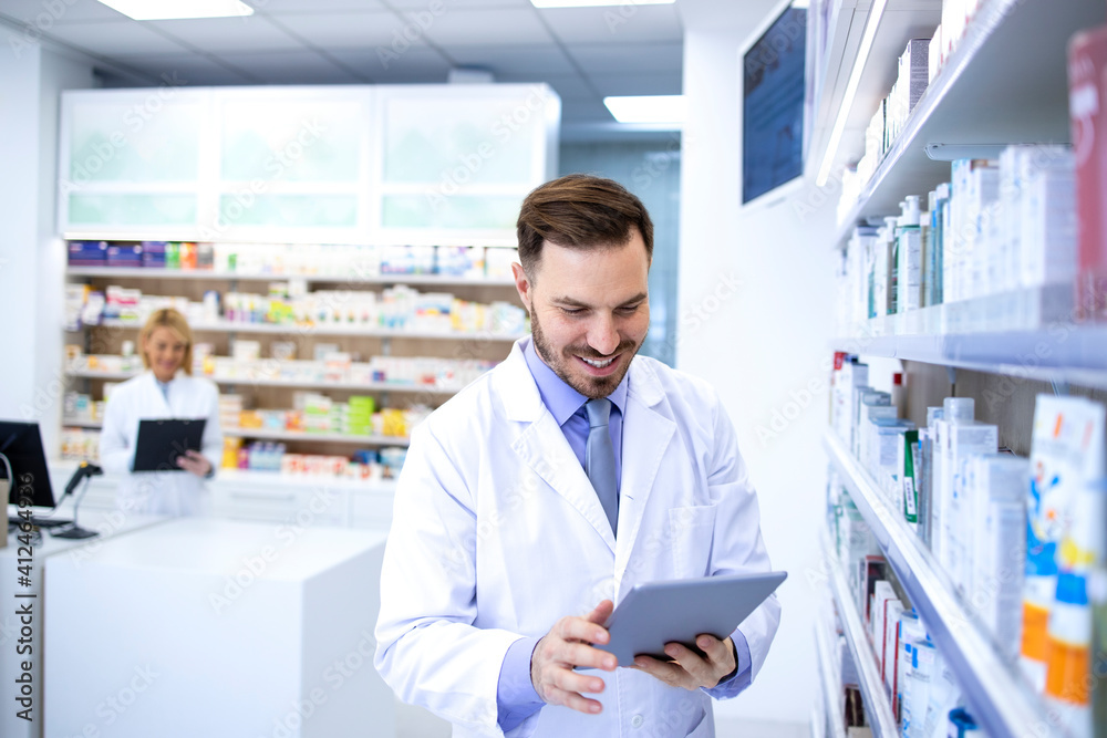 Handsome male pharmacist in white coat working in pharmacy store or drugstore. Checking medicines on his tablet computer. Healthcare and apothecary. Handsome male pharmacist in white coat working in pharmacy store or drugstore. Checking medicines on his tablet computer. Healthcare and apothecary.