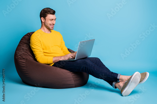 Full size profile photo of cheerful handsome guy sitting big soft chair use netbook isolated on blue color background