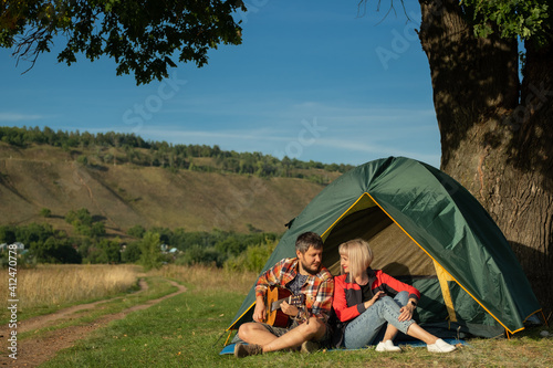Man and woman sing a song with guitar sitting at tent. Family is on camping trip.