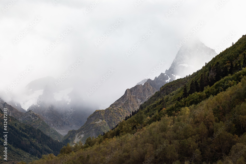 Snow capped mountain range in the clouds. Dense forest on the mountain slopes Caucasus, Russia Snow capped mountain range in the clouds. Dense forest on the mountain slopes Caucasus, Russia