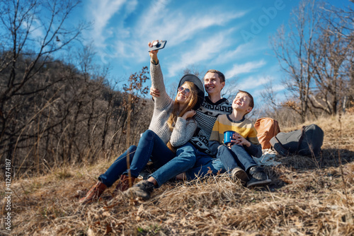 Beautiful young family with kid son sitting in a park making selfie on a smartphone in a spring sunny forest