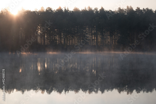 Cold summer morning in the forest with lake, forest reflection and mist on the water surface.