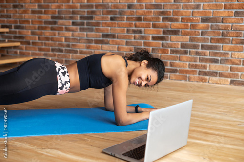 Training at home concept. Fit African-American woman doing plank exercise, watching online tutorial on laptop, exercising in modern loft apartment