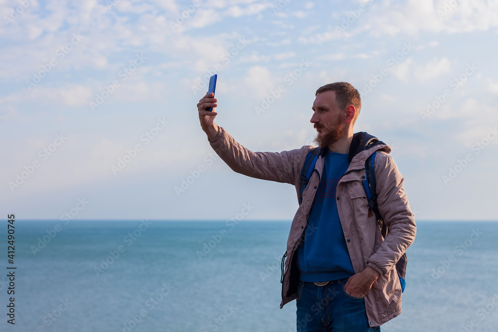 Bearded man in raincoat with backpack on sea landscape with blue cloudy sky background with smartphone in hand taking picture. Guy photographing nature on cell phone, active lifestyle travel concept. Bearded man in raincoat with backpack on sea landscape with blue cloudy sky background with smartphone in hand taking picture. Guy photographing nature on cell phone, active lifestyle travel concept.