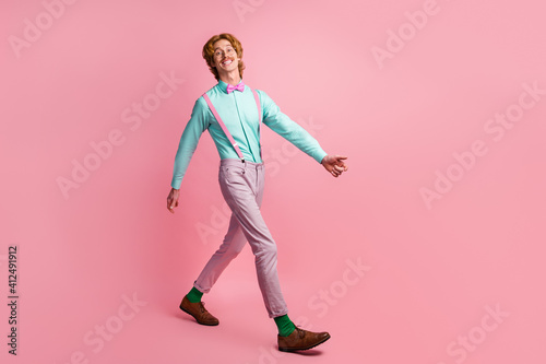 Full length body size photo smiling man wearing pants suspenders teal shirt walking forward isolated pastel pink color background