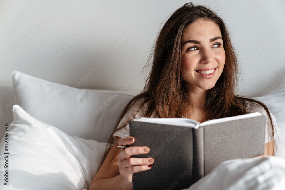 Joyful caucasian woman smiling and reading book while resting in bed