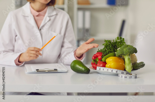 Cropped image female nutritionist points to a plate of fresh vegetables and a tape measure on her desk. Concept of a healthy lifestyle, body care and weight loss. Selective focus. Blurred background.