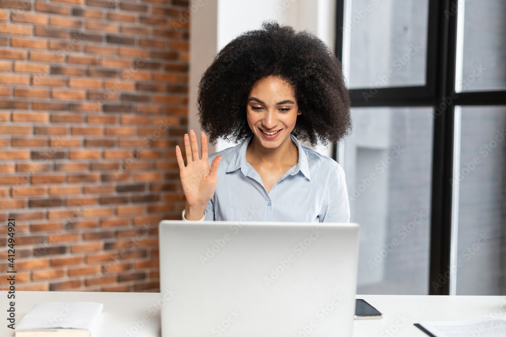 Beautiful young American woman with curly hairstyle sitting at the laptop, chatting with friends, talking about the news, greeting the colleagues, working from home Beautiful young American woman with curly hairstyle sitting at the laptop, chatting with friends, talking about the news, greeting the colleagues, working from home