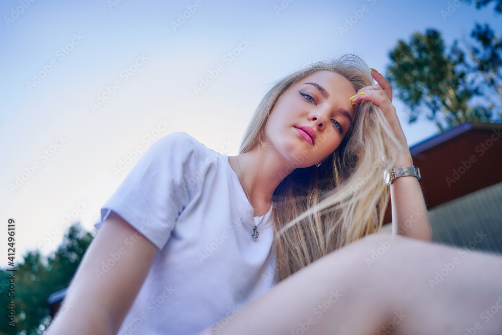 Charming teenage girl in a white T-shirt against a blue sky sits in a park and straightens her hair. View from below. Charming teenage girl in a white T-shirt against a blue sky sits in a park and straightens her hair. View from below.