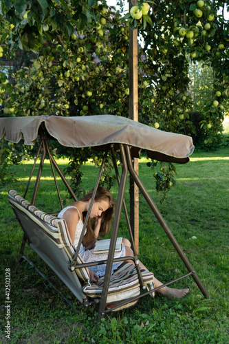 Teenage girl reads a book sitting on the garden swing in summer garden under an apple tree