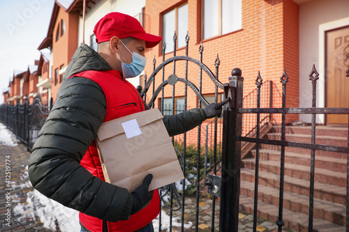 Courier in medical mask holding paper bag with takeaway food and ringing gate bell outdoors. Delivery service during quarantine due to Covid-19 outbreak